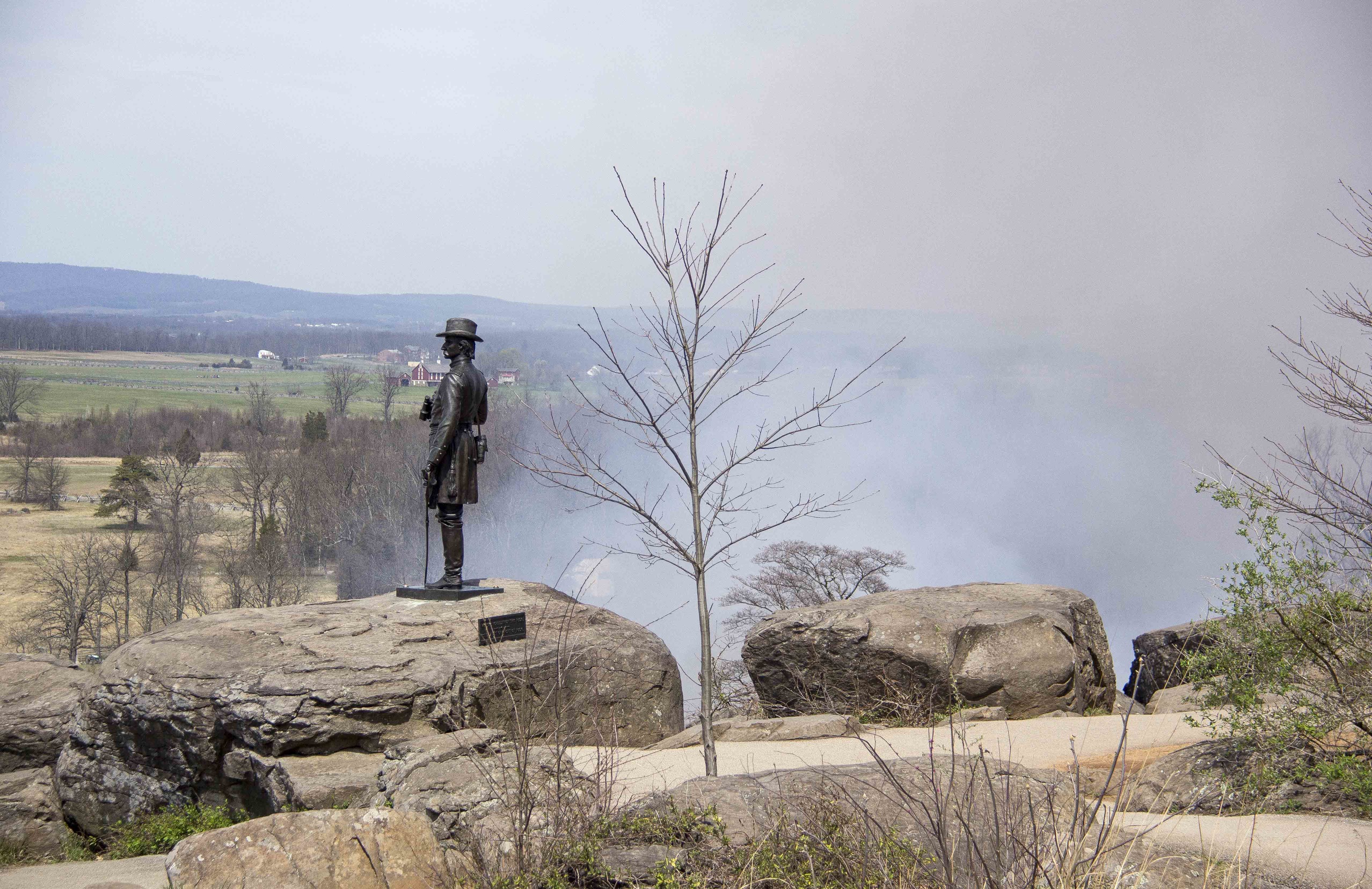 The monument to Brigadier General Gouverneur K. Warren stands atop the summit of Little Round Top as the smoke from the prescribed fire slowly creeps around the hill. There are many large boulders, some brush, and one tree in the picture.