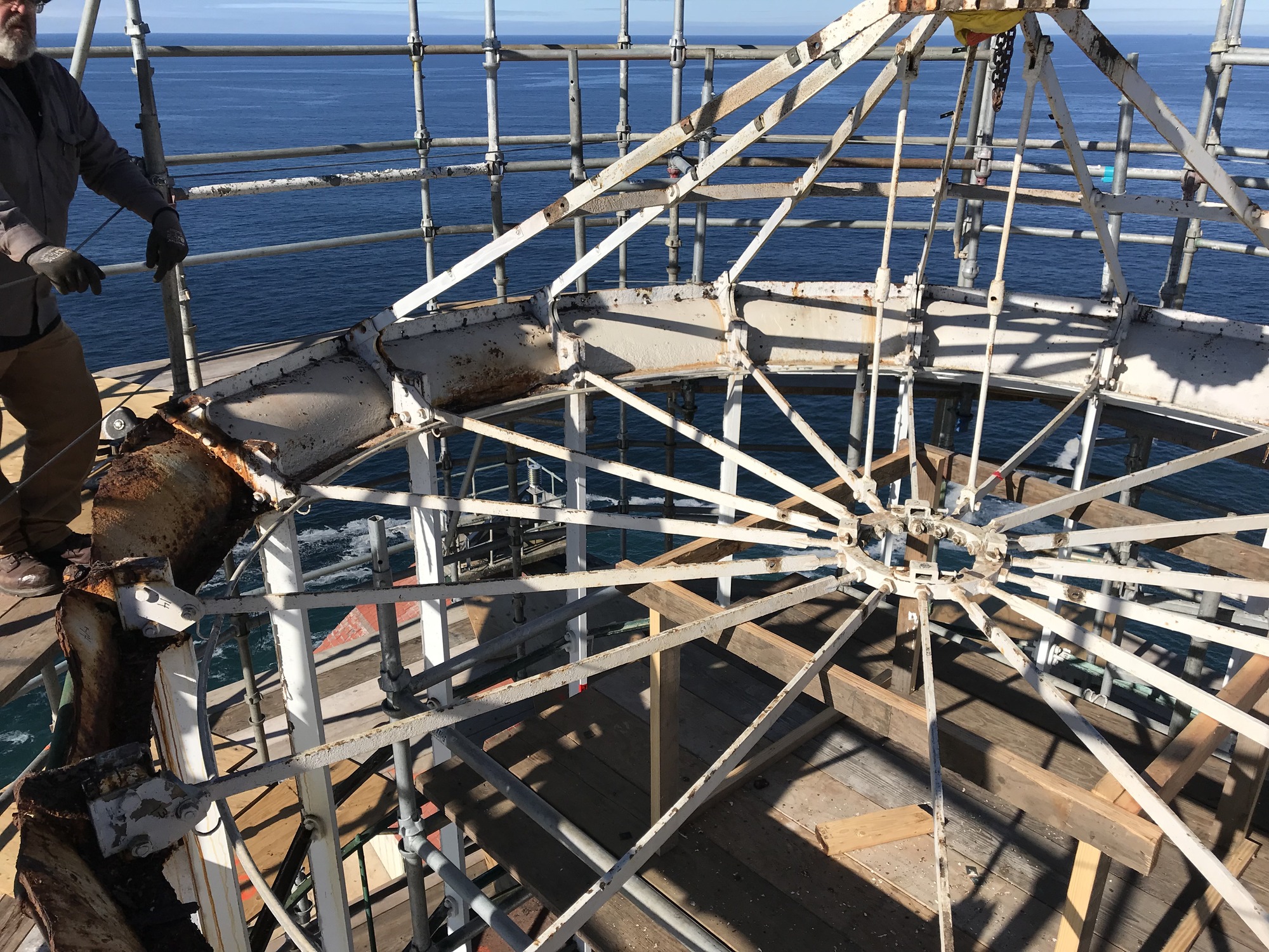 Six out of an original sixteen white diagonal cast iron rafters descend from a central peak to a circular green ring at the top of the roofless lighthouse, which is surrounded by scaffolding. Sixteen white horizontal bars radiate from the center of the tower below the rafters.