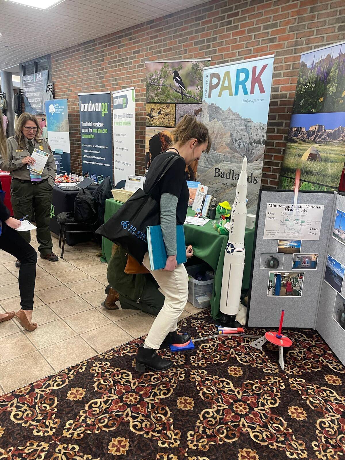 Woman in black shirt and white pants steps on a stomp rocket launcher in a hotel lobby beside a white model missile. 