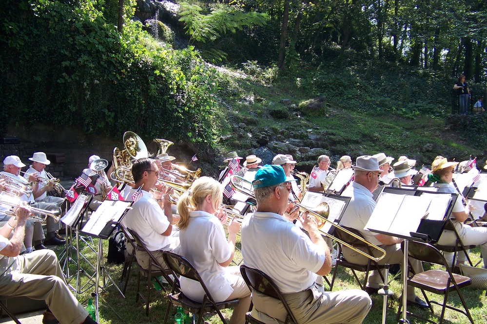 Side view of community band, showing members seated in rows with music stands in front of each. Back row shows trumpets, next row shows trombones and French horns, and other instruments toward the front rows. Behind the band you can see the hot spring water cascade.