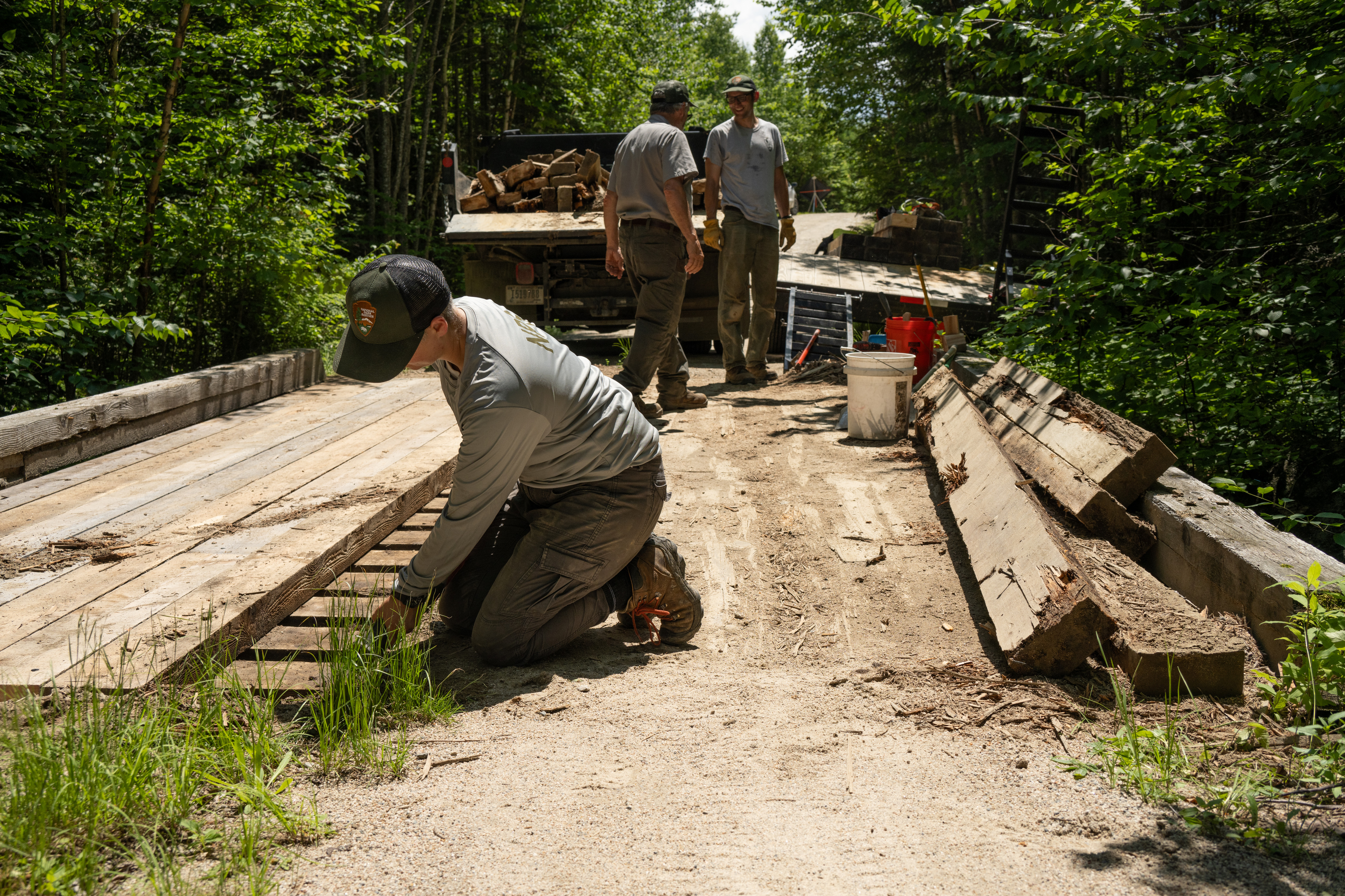 A worker kneels on a gravel bridge and lays down planks of wood. In the background two more workers stand by a tuck filled with wood.