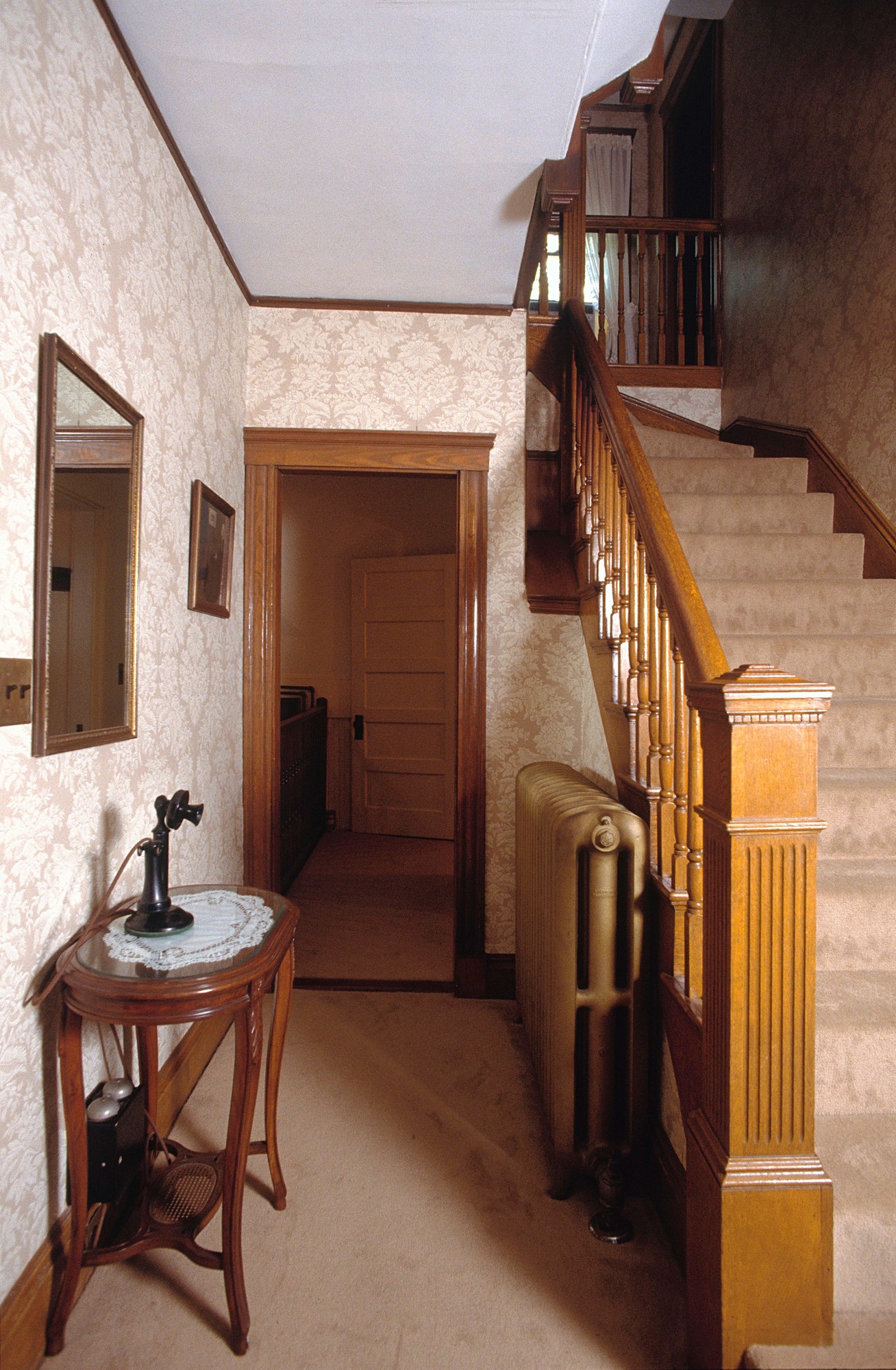 Hallway with staircase on right. Tan damask pattern wallpaper, wooden trim, banister, and newel post. A black candlestick style phone stands on a hall table in front of mirror.