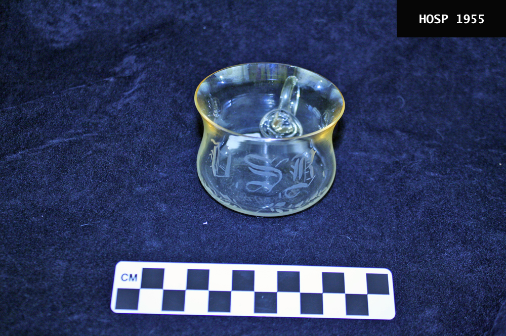 Clear glass cup slightly flared at top and base with loop handle of glass, engraved in front and on bottom. Pale gold patina said to have been acquired from immersion in hot spring water while the purchaser was bathing. Bathhouses sold these to their customers as souvenirs around 1900. This one was sold at the Superior Bathhouse (probably the first one).