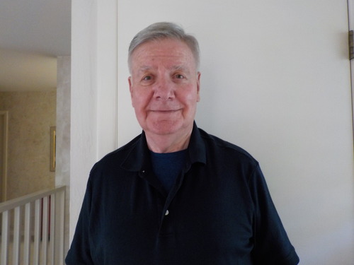 Image of Douglas Bereuter in his home. Man with grey hair and navy shirt standing by white wall.