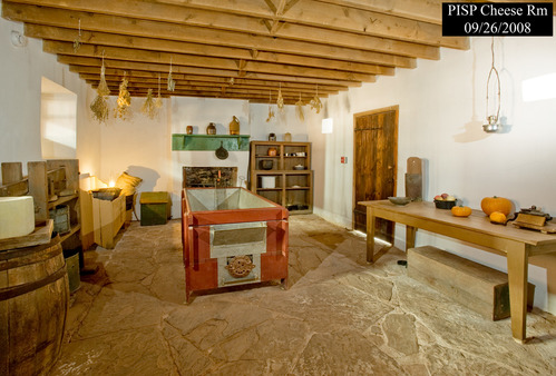 A cheese making double boiler vat sits in the center of a room with a flagstone floor. Around the edges of the room are tables, presses, and other equipment needed to manufacture cheese.