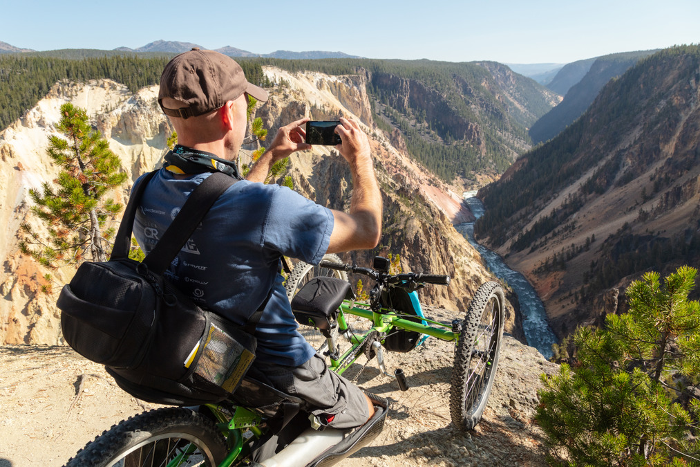 Man in three wheeled chair sits on the rim of a deep canyon and takes a photo of it.