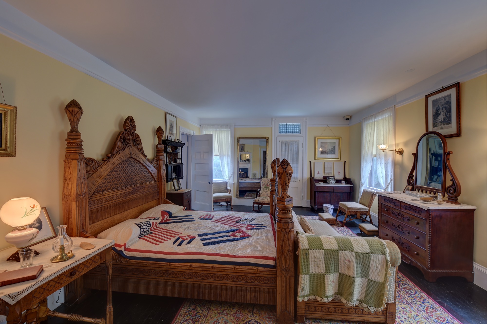 A yellow bedroom with a grand, ornately carved wooden double bed in the center. There is a red, white, and blue quilt on the bed. There are also two nightstands, a chaise lounge, a dresser with a mirror, a washstand, a tall mirror, and three chairs. 