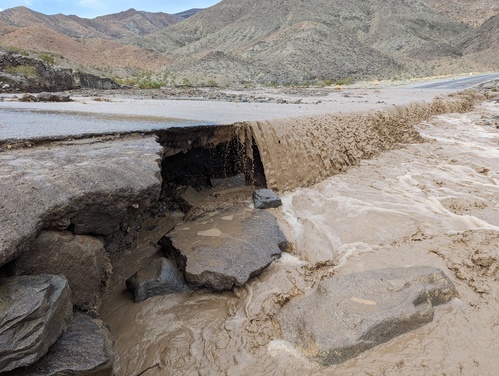 Side view of muddy flood water flowing over a paved two-lane road and undercutting it on the downhill side, with desert mountains in the background.