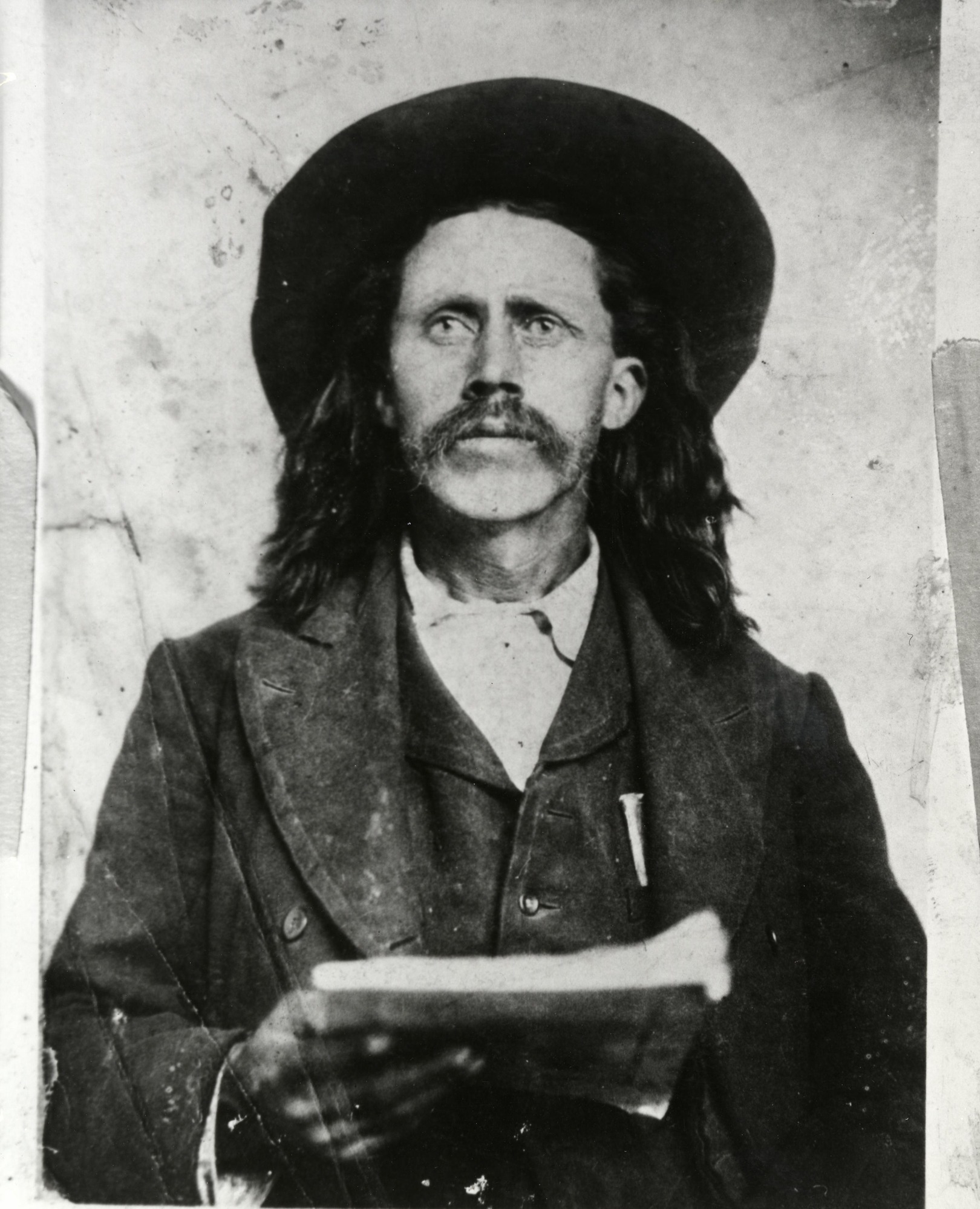 A white man with longer hair wearing a dark suit holding papers in his hand.