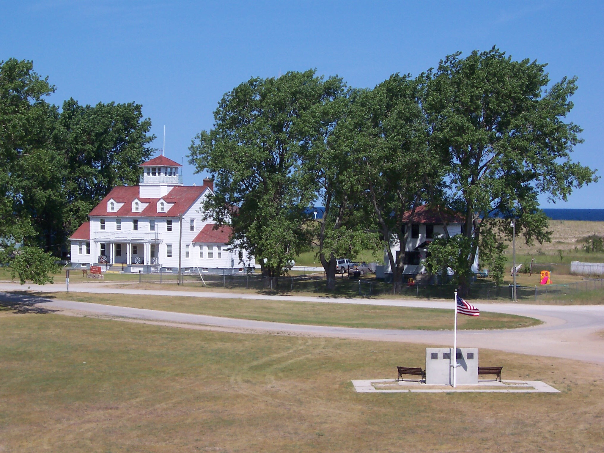 Three story white wooden Coast Guard building, two story white house hidden behind large poplar trees, and small memorial to fishermen.