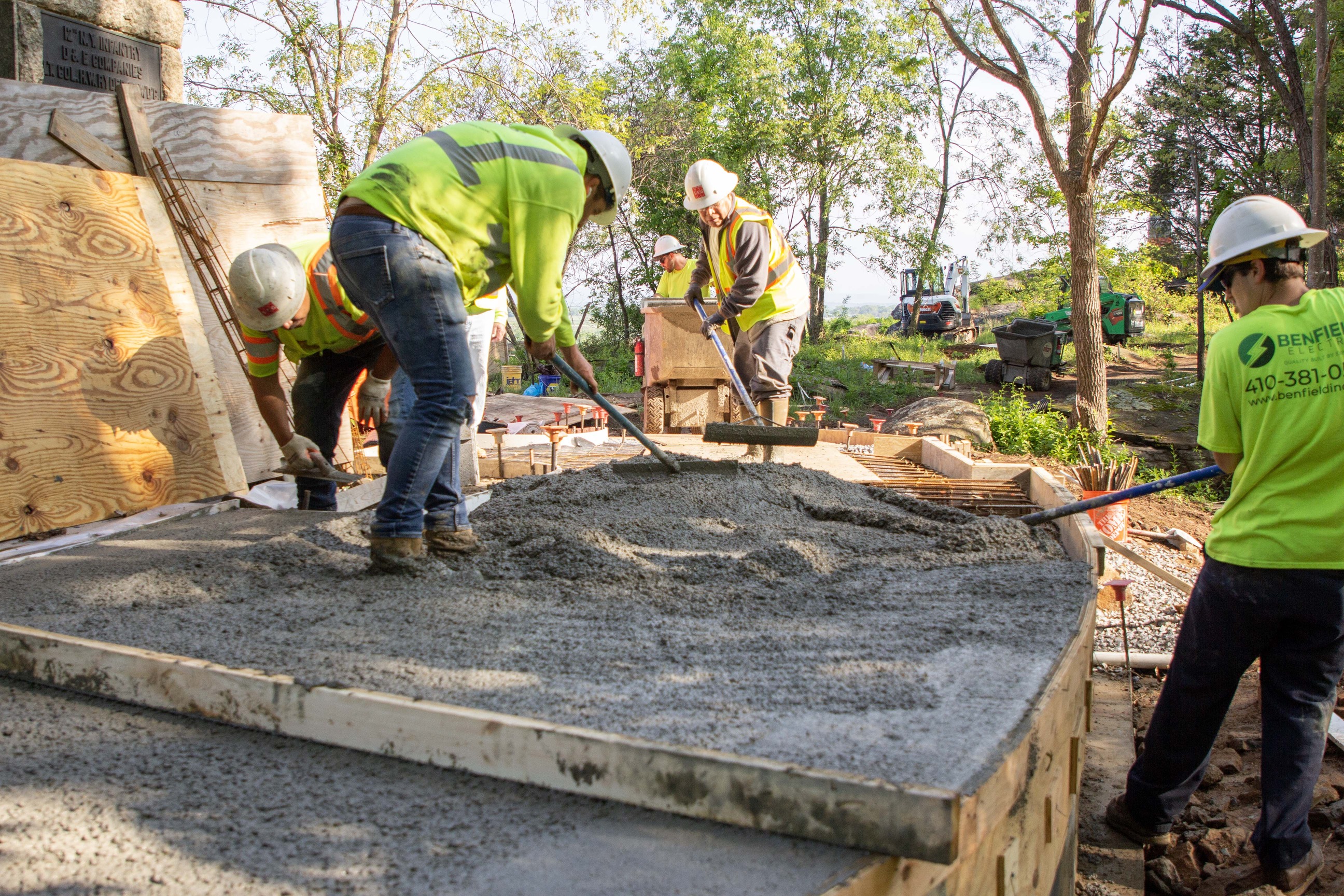 Construction workers stand in freshly poured concrete to smooth it out next to a monument, with trees and construction machinery in the background.