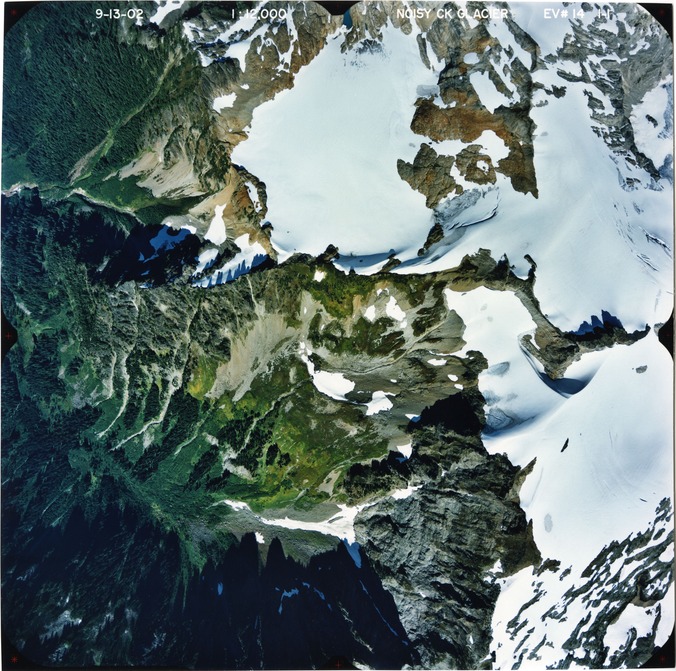 Color aerial photograph of glacier on mountain peak, surrounded by forested valleys.