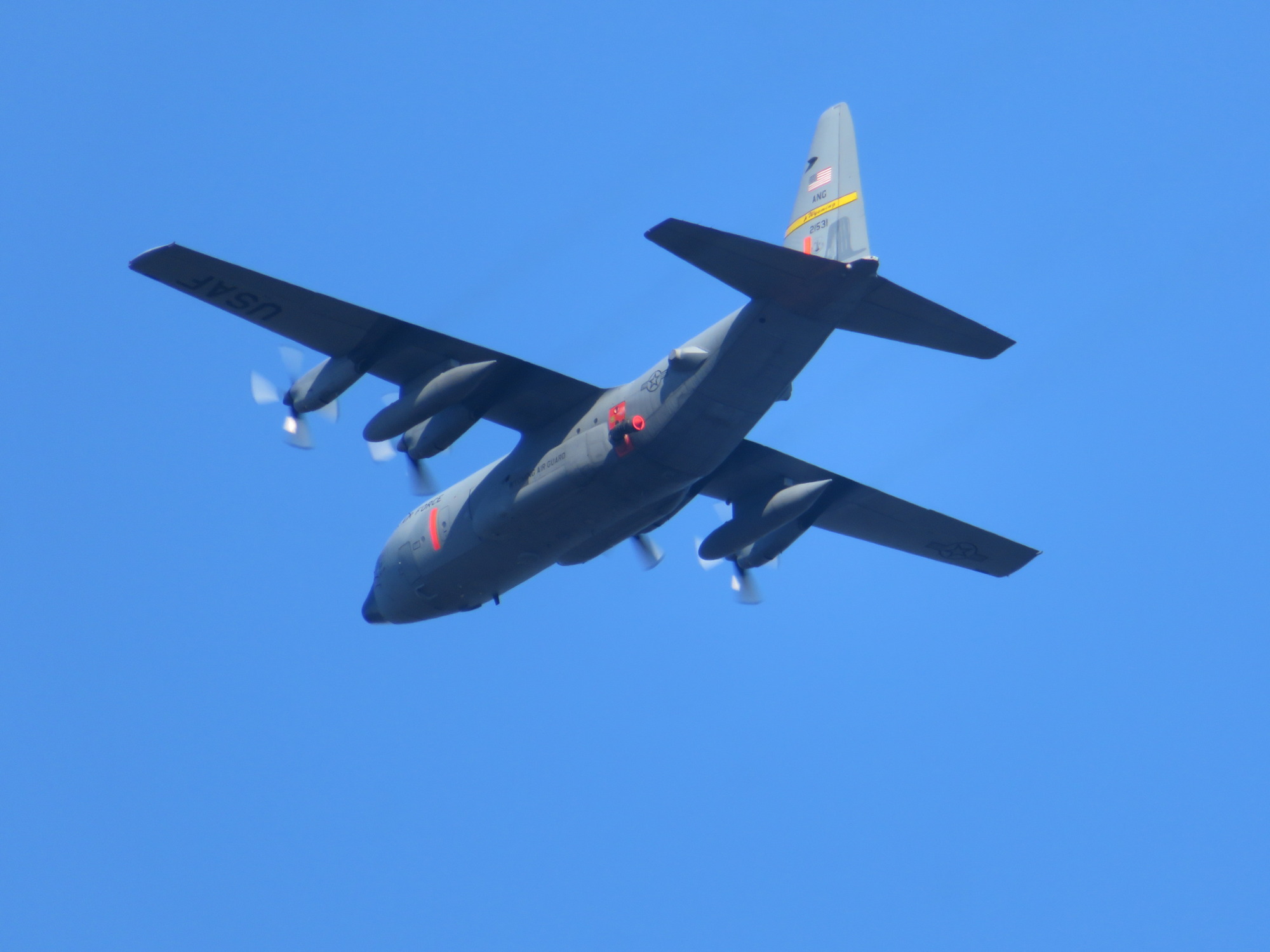 A large gray four-engine turboprop military transport aircraft flies through blue skies.