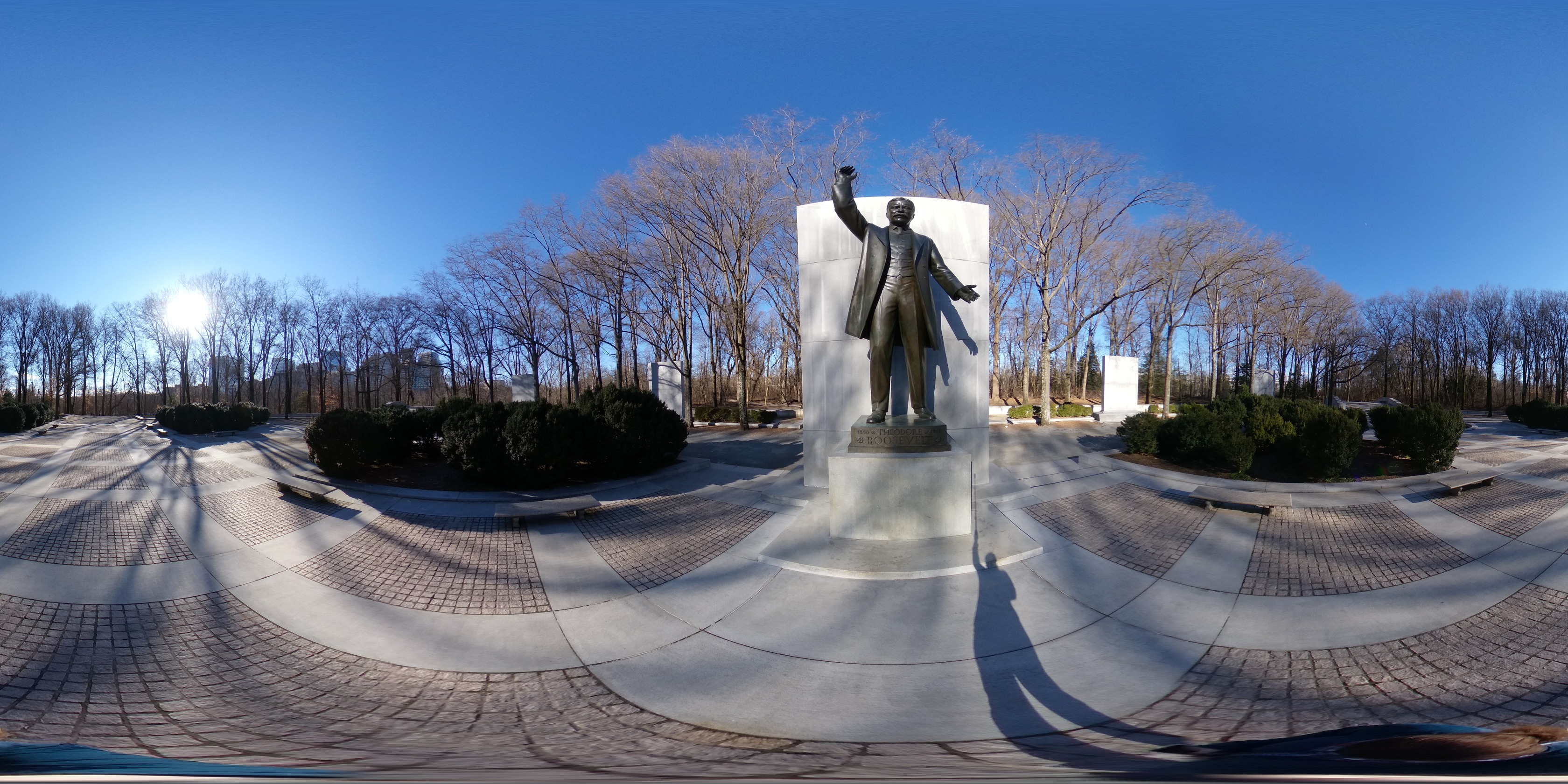 Spherical image of a memorial plaza surrounded with woods. The plaza include a large statue of Theodore Roosevelt, two large fountains ponds, and two bridges over pools.