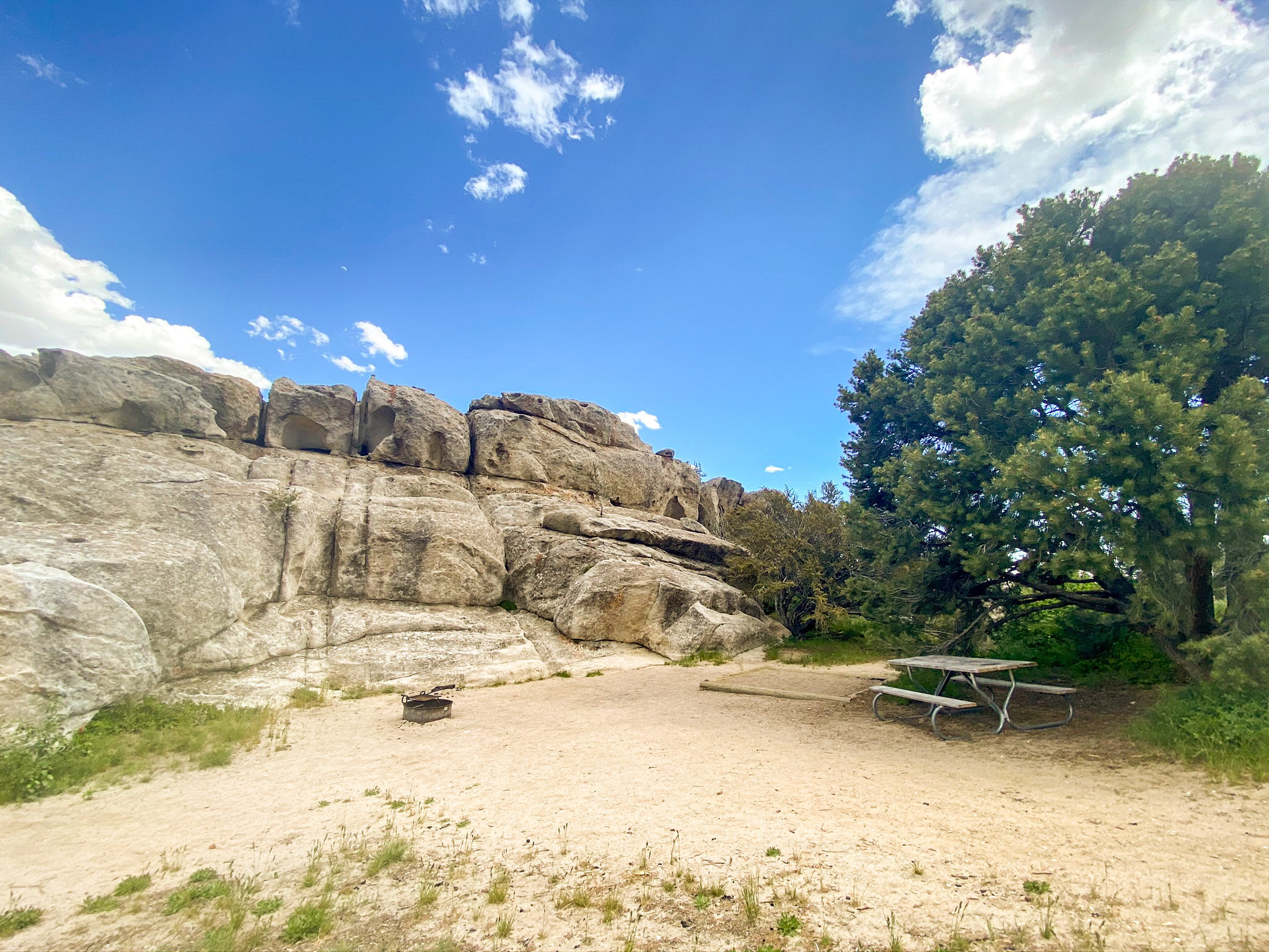 Open sandy area with a large pine tree, fire ring, tent pad, and picnic table at the base of a granite formation.