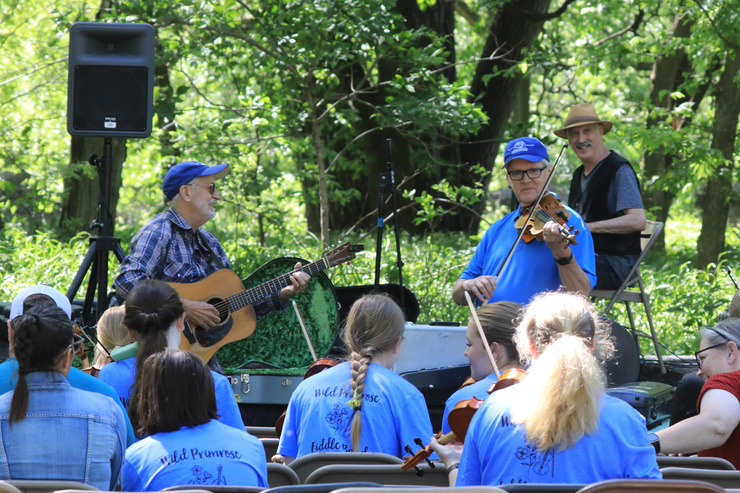 Two men stand in front of an outdoor class. One plays the fiddle. The other plays guitar. Fiddles can be seen in the hands of several audience participants.