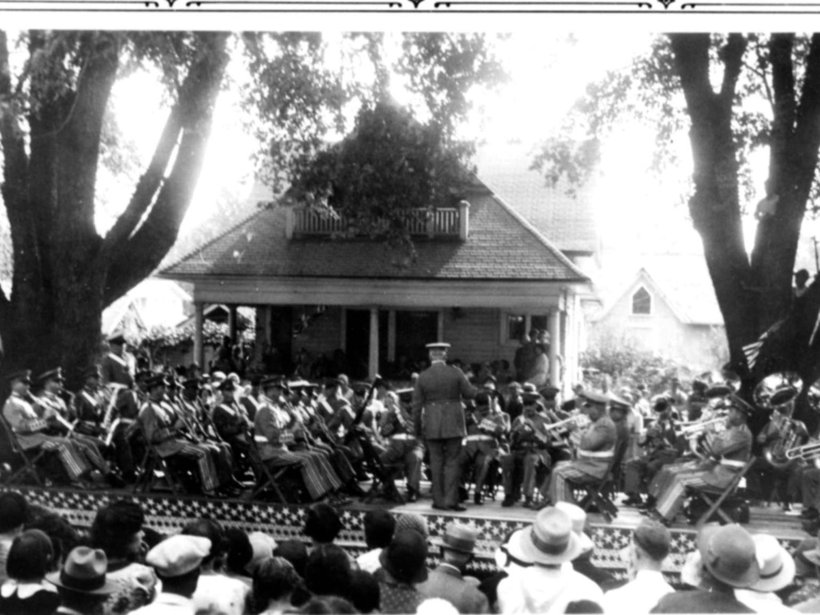 A crowd gathers to hear an Army band playing in front of a house with a porch.