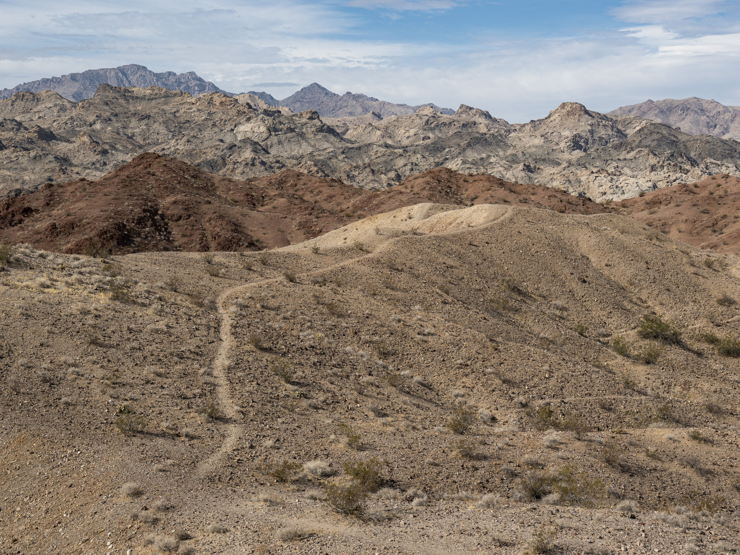 narrow trail leading through center, hills in foreground, rugged mountains and sky in distance