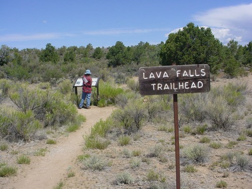 A brown wooden sign reads "Lava Falls Trailhead" in white letters.  A dirt trail is immediately left of the sign.  A hiker stands at a wayside sign.