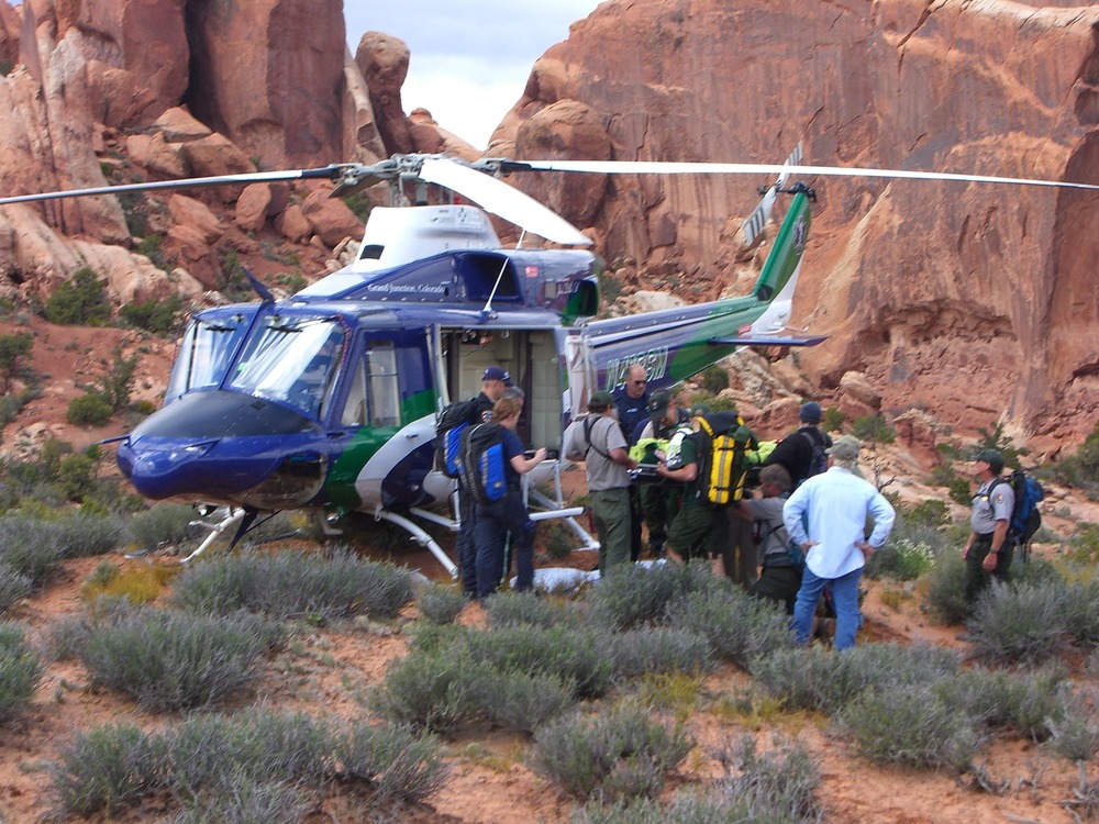 A group of people gather around a patient on a wheeled medical litter next to a helicopter.  Large sandstone cliffs tower behind.