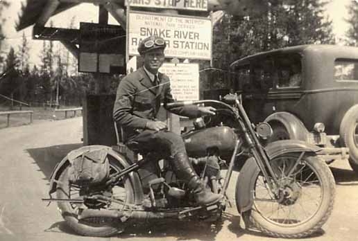 Archival photograph of an NPS park ranger on a motorcycle.  The ranger wears full riding garb, including goggles on his forehead.  A Model T is parked behind the ranger and motorcycle.