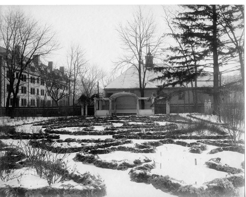 Black and white photograph of formal garden covered in snow. 1860's dorm building visible in background.