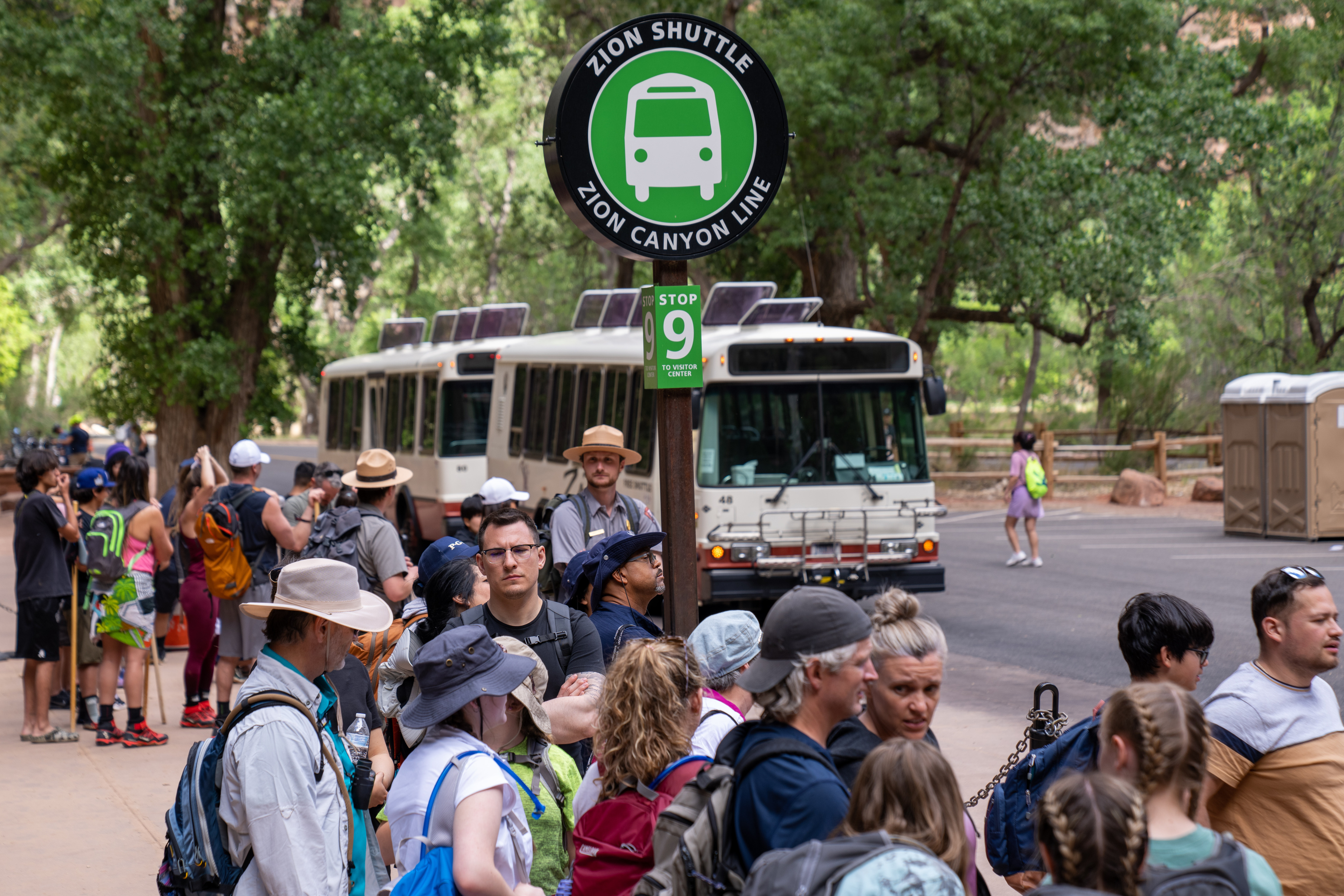 Line of visitors waits to board shuttles at Temple of Sinawava in Zion National Park. Two uniformed NPS rangers standby to direct traffic.