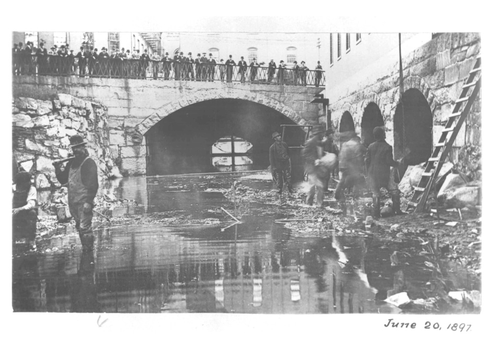A group of men, who are covered in dirt, pose for a picture while rich guys in suits lok