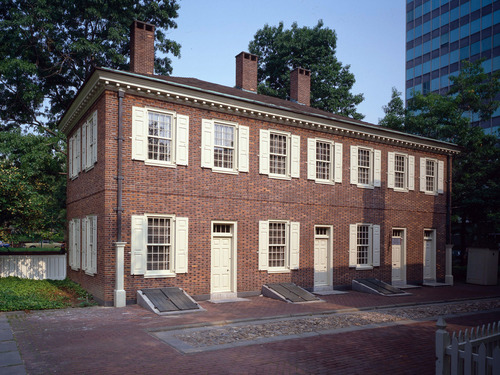 Color photo of a two story red brick building, rectangular in shape, with rows of windows flanked by cream shutters.