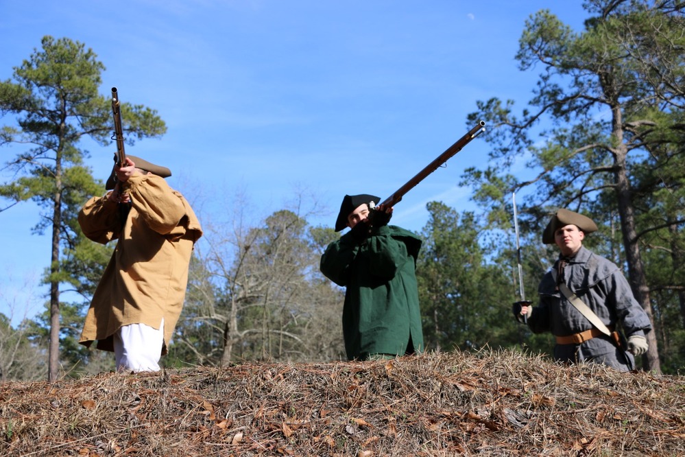 Two Patriots stand poised to fire muskets, with a third Patriot wielding a sword.