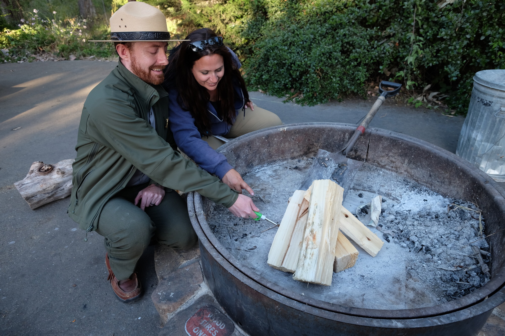 A Park Ranger and a woman crouch near a fire pit, ready to light logs on fire.