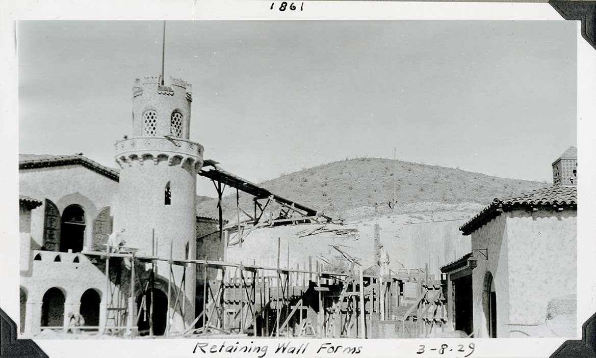 This is an historic black and white photograph from the Scotty's Castle Historic Photograph Collection, Death Valley National Park of wood ramp in foreground. Three men working in different areas. Two story building with round flag tower on left. Hill in background. Smaller building with Spanish tile roof on right. Inscriptions in black ink along upper and lower border.
