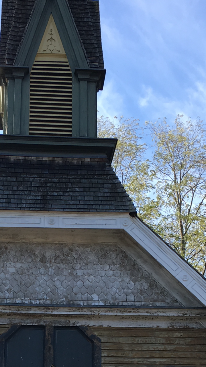 left half of the image shows the green church steeple with yellow slats and ornamental design in front of the belfry. Weathered shingles top the steeple and the church roof around it. right side of the image is blue sky with  wispy white clouds.