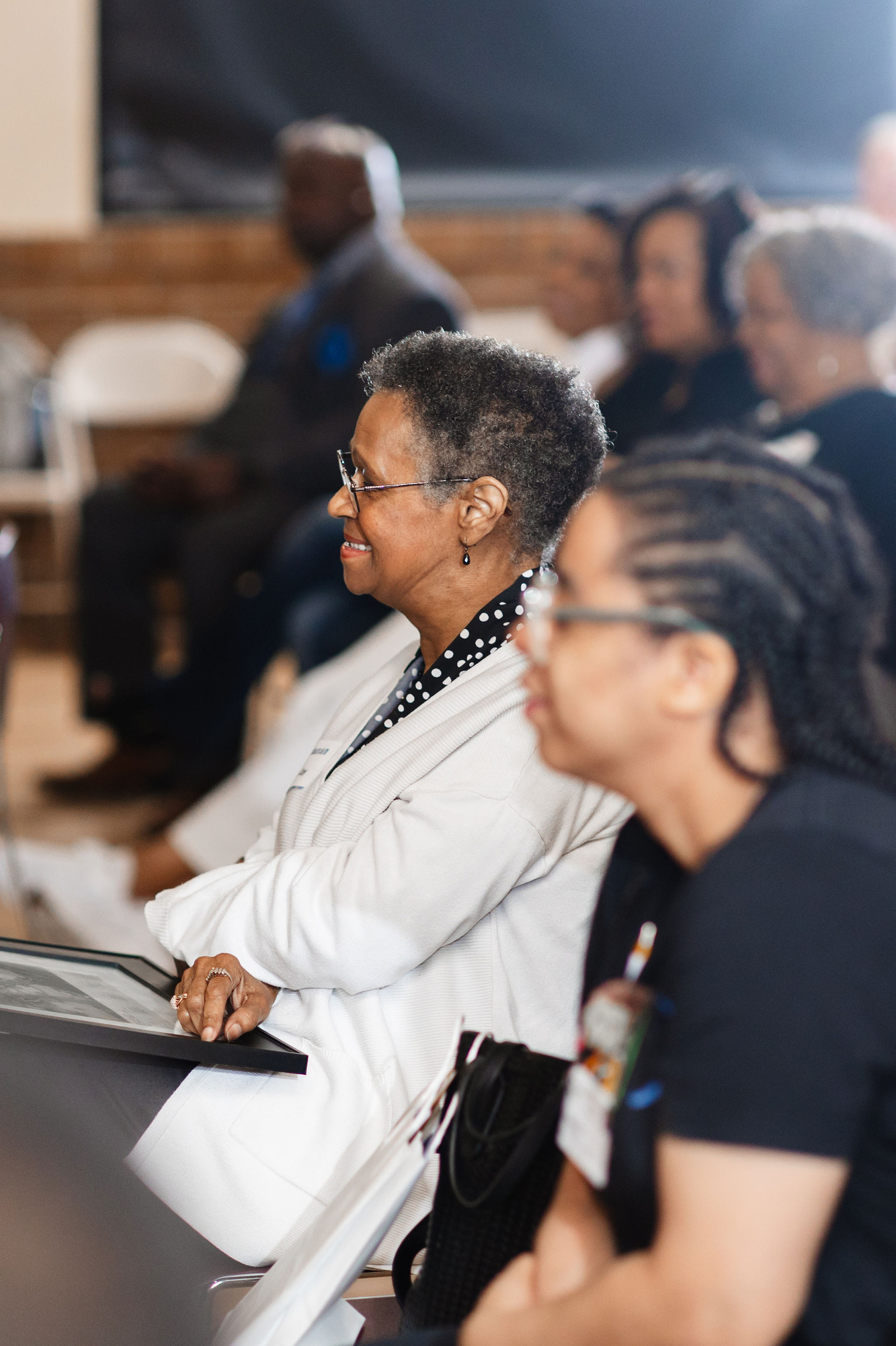 African American adults sit in maroon plastic chairs in a room with hardwood floors the photo focuses on an older african american woman in a white blazer smiling brightly