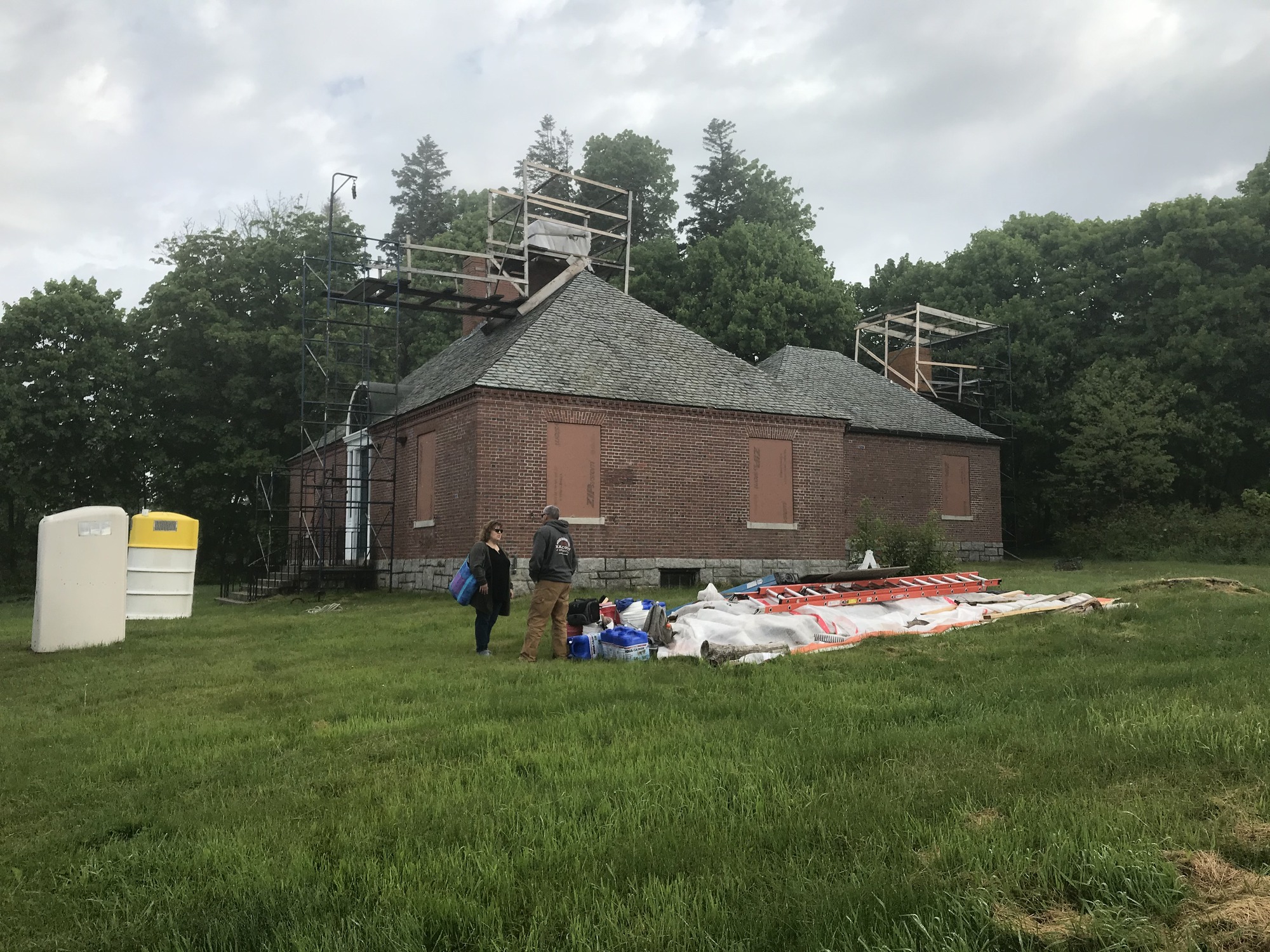 Two large structures protect the chimney and shutters are closed, portable toilets are outside the building, and building materials on the grass.