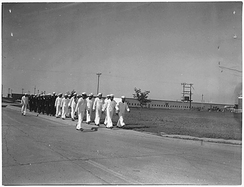 Black and white photo of men in sailor uniforms marching down a large empty road.