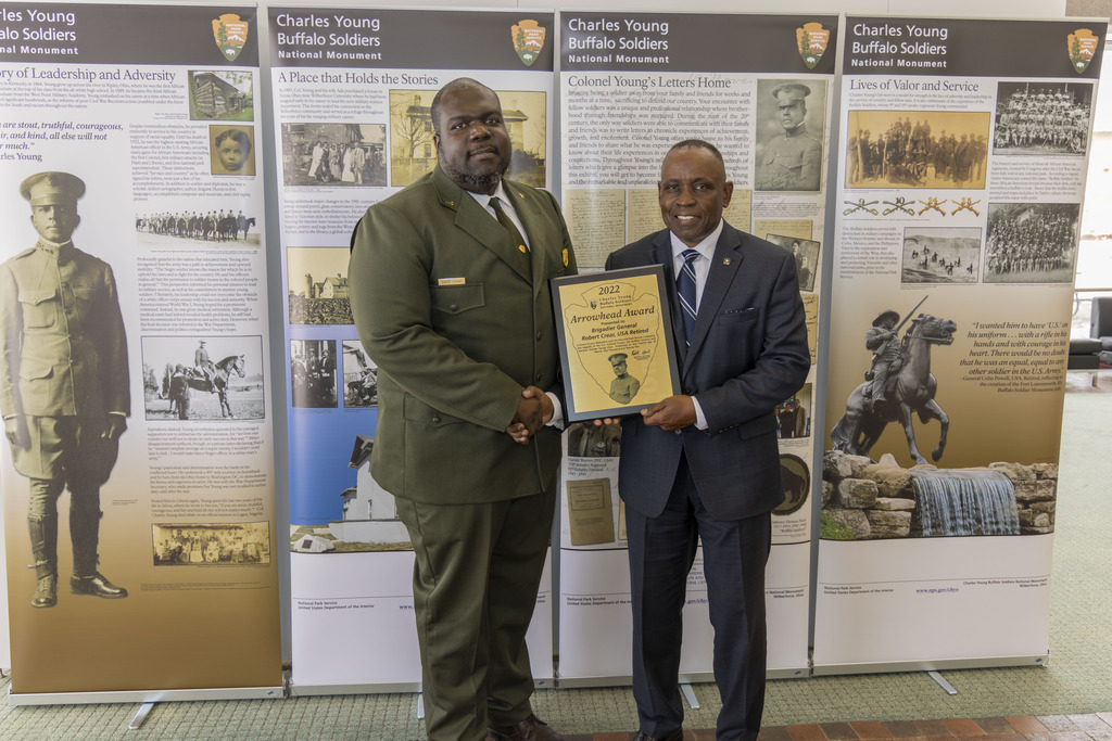 A park ranger in a green suit and tie stands next to a man in a dark-colored suit as they both shake hands. The man on the right is holding a wooden plaque with a bronze plate attached to the front of it.