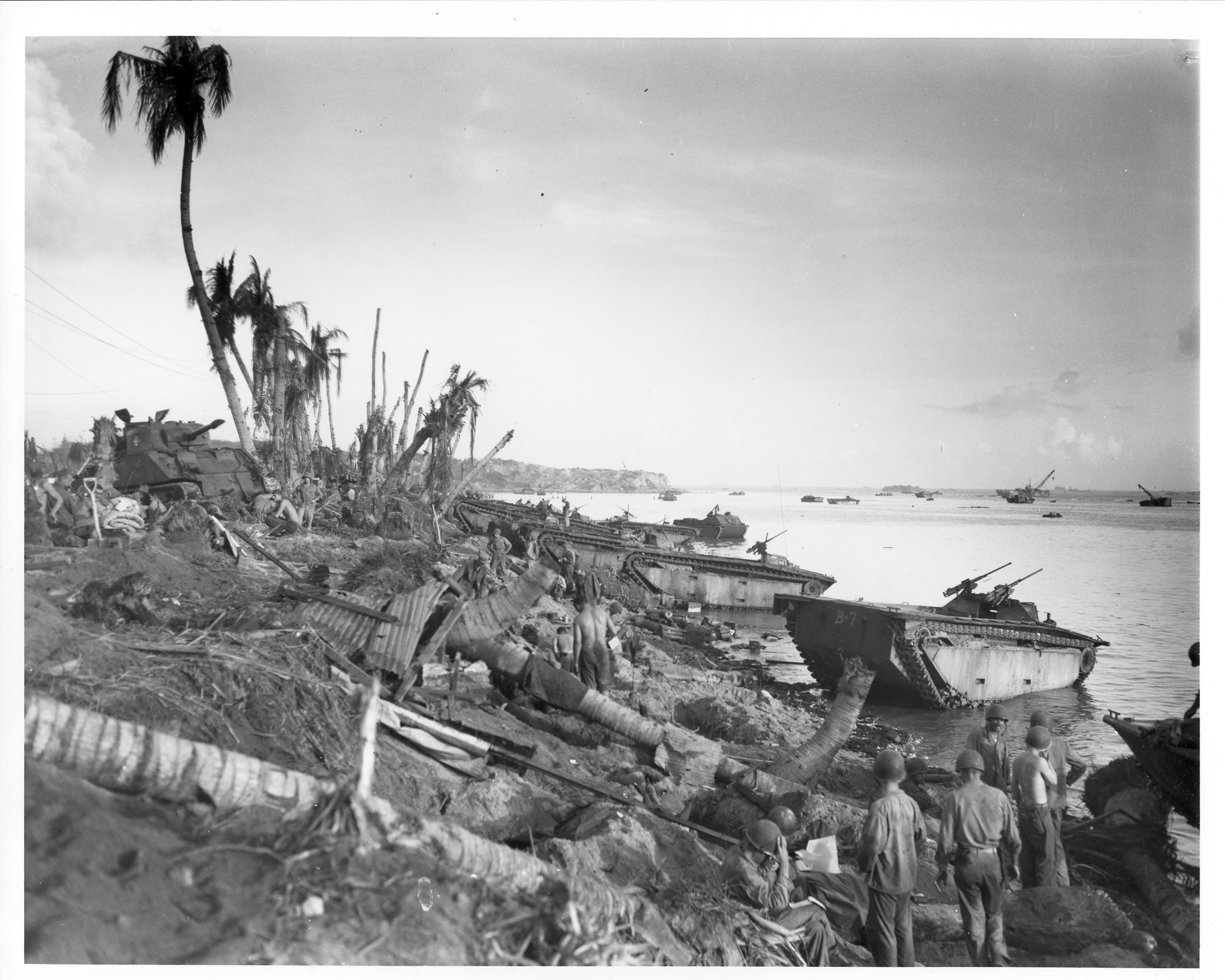 Soldiers walk over a beach full of blown down threes and landing vehicles.