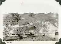 This is an historic black and white photograph from the Scotty's Castle Historic Photograph Collection, Death Valley National Park of a large l-shaped building with a flat roof setting along the length of a wash at base of barren desert mountains. Another building at right edge interrupted by plane prop. Number in black ink in lower left corner.