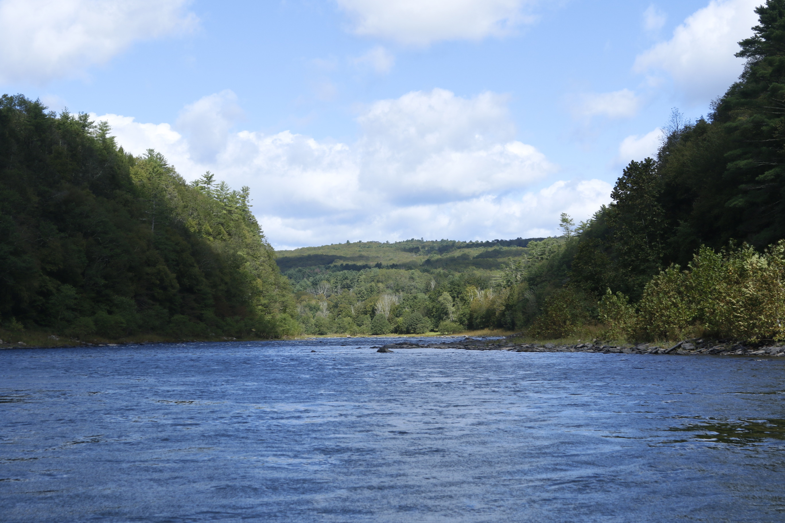 View on blue river, reflecting pale blue sky with white clouds. River is flanked on both sides by large , green forested hills.
