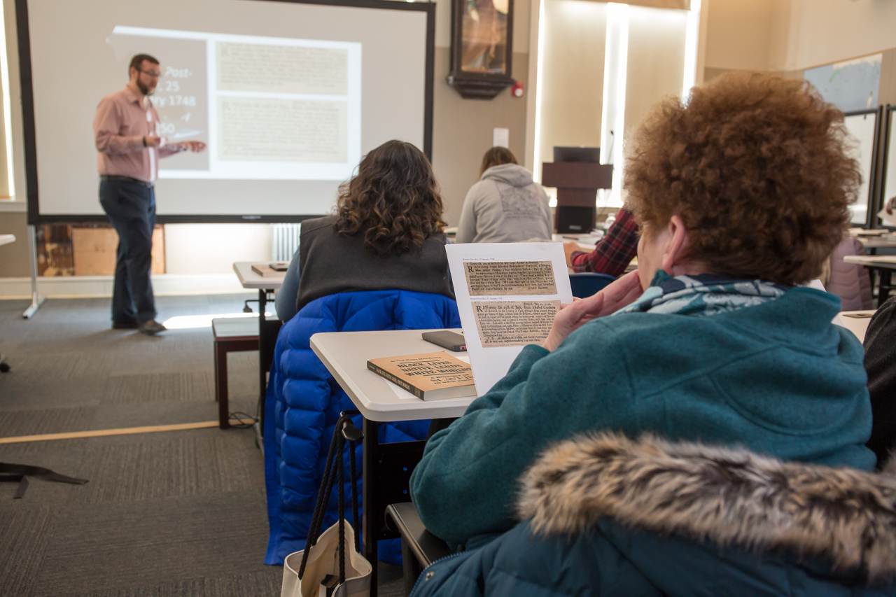 Woman with her back to the camera sits at a table and looks at a piece of paper she is holding.  More people sitting at tables are visible but out of focus. A man stands at the front of the room speaking. 