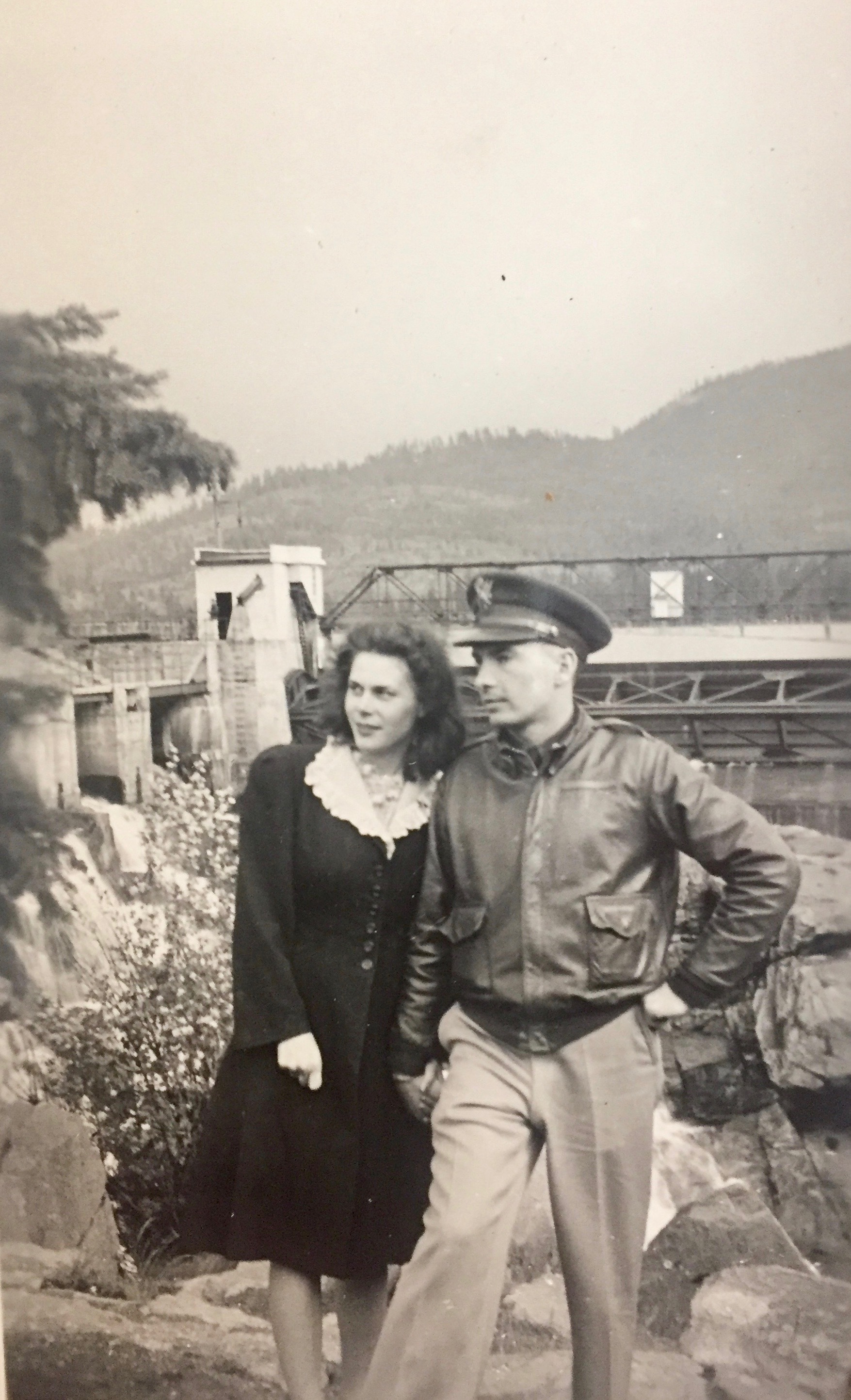 Man in partial uniform and woman in dress pose in front of dam. 