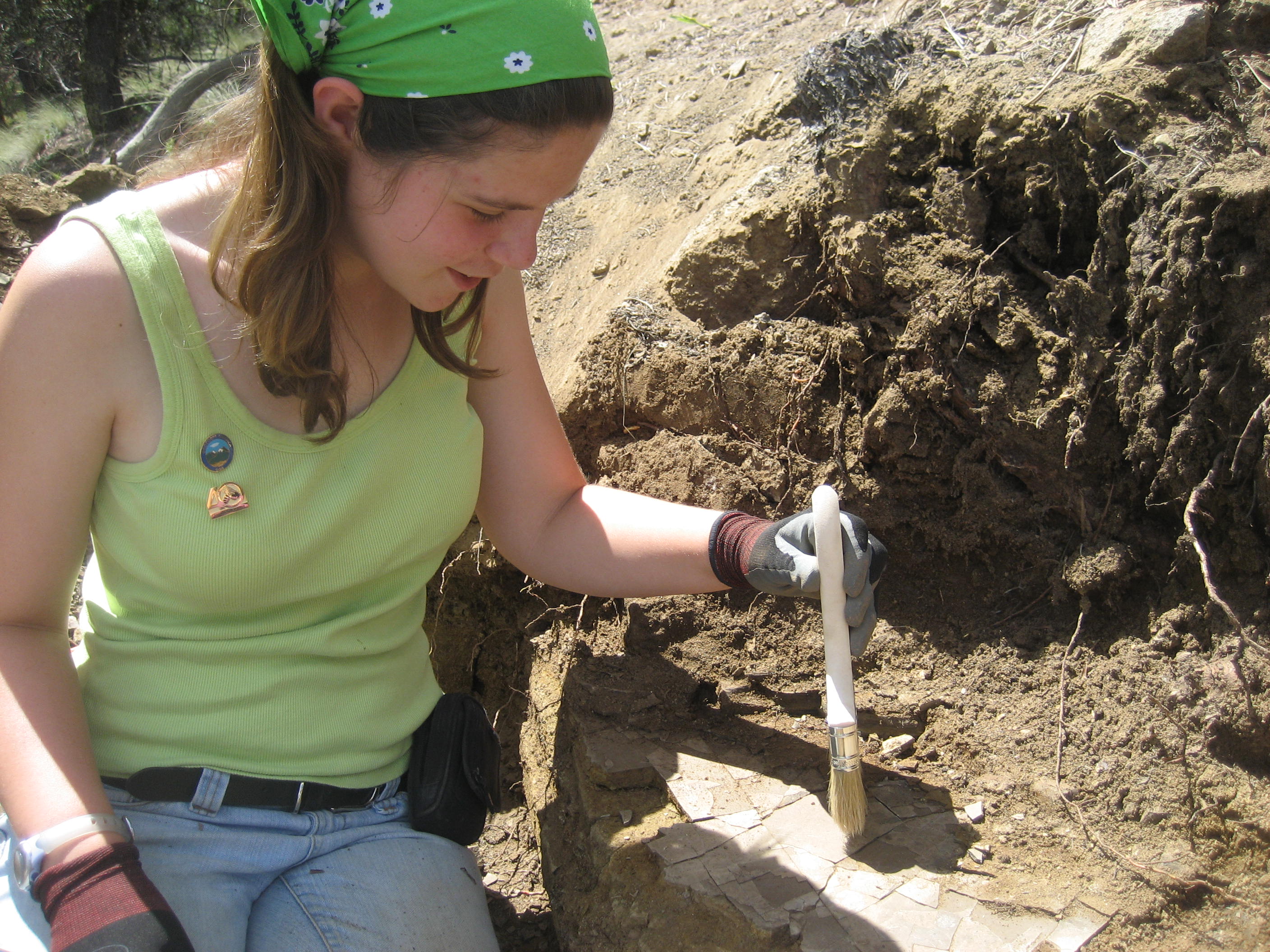 a young woman sits on a dirt edge and brushes off dirt from some rocks. 