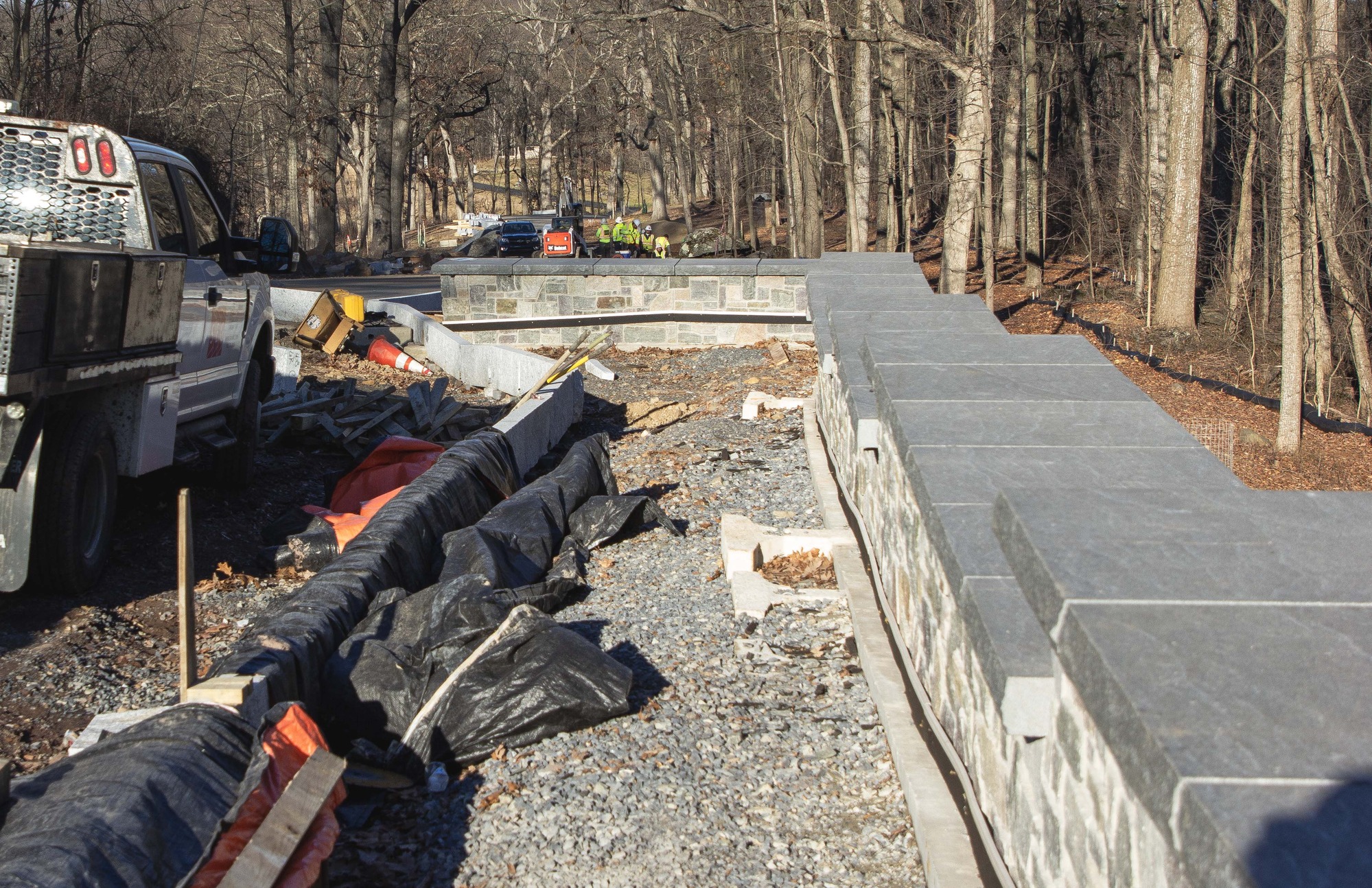 A gravel path is outlined by a rock wall and stone curbing. Trees line the upper portion of the photograph. A construction vehicle appears in the left side of the photograph. A group of construction workers in yellow jackets stand together in the background of the photograph. 
