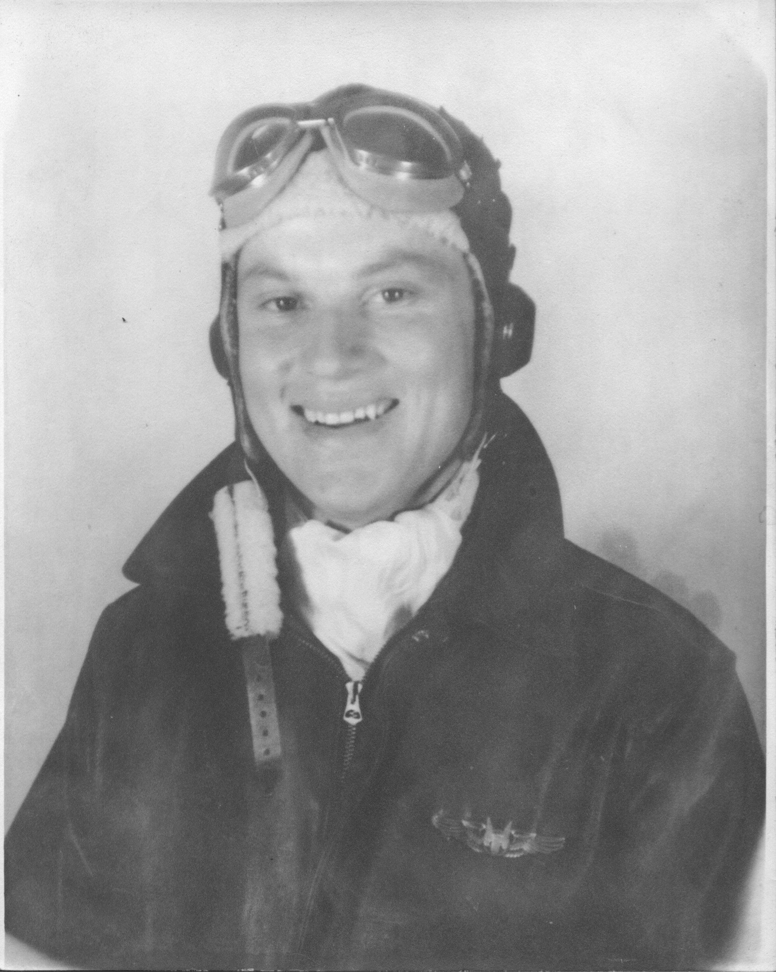 Black and white headshot of white man smiling in a leather jacket with a white scarf and aviation goggles and leather helmet. 