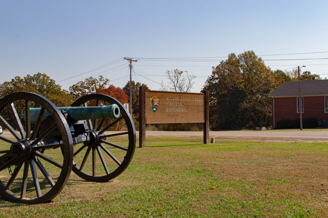 A full color photograph of a grassy field with fall foliage in the background. In the left side of the frame is a Civil War-era cannon. On the right side of the frame is a red building with a gray roof. Center of the image, a little farther back, is a brown wooden sign with the National Park Service arrowhead logo and text that reads "Brices Cross Roads National Battlefield".