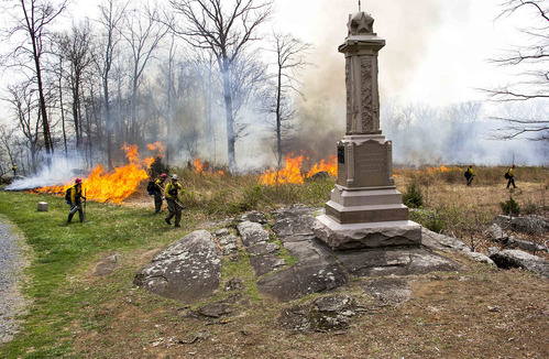 Several crew members carefully monitor the flames as the prescribed fire continues around the 29th Pennsylvania Infantry monument. The crew members are spread out in the background. The monument is in the foreground. The flames are in the background.