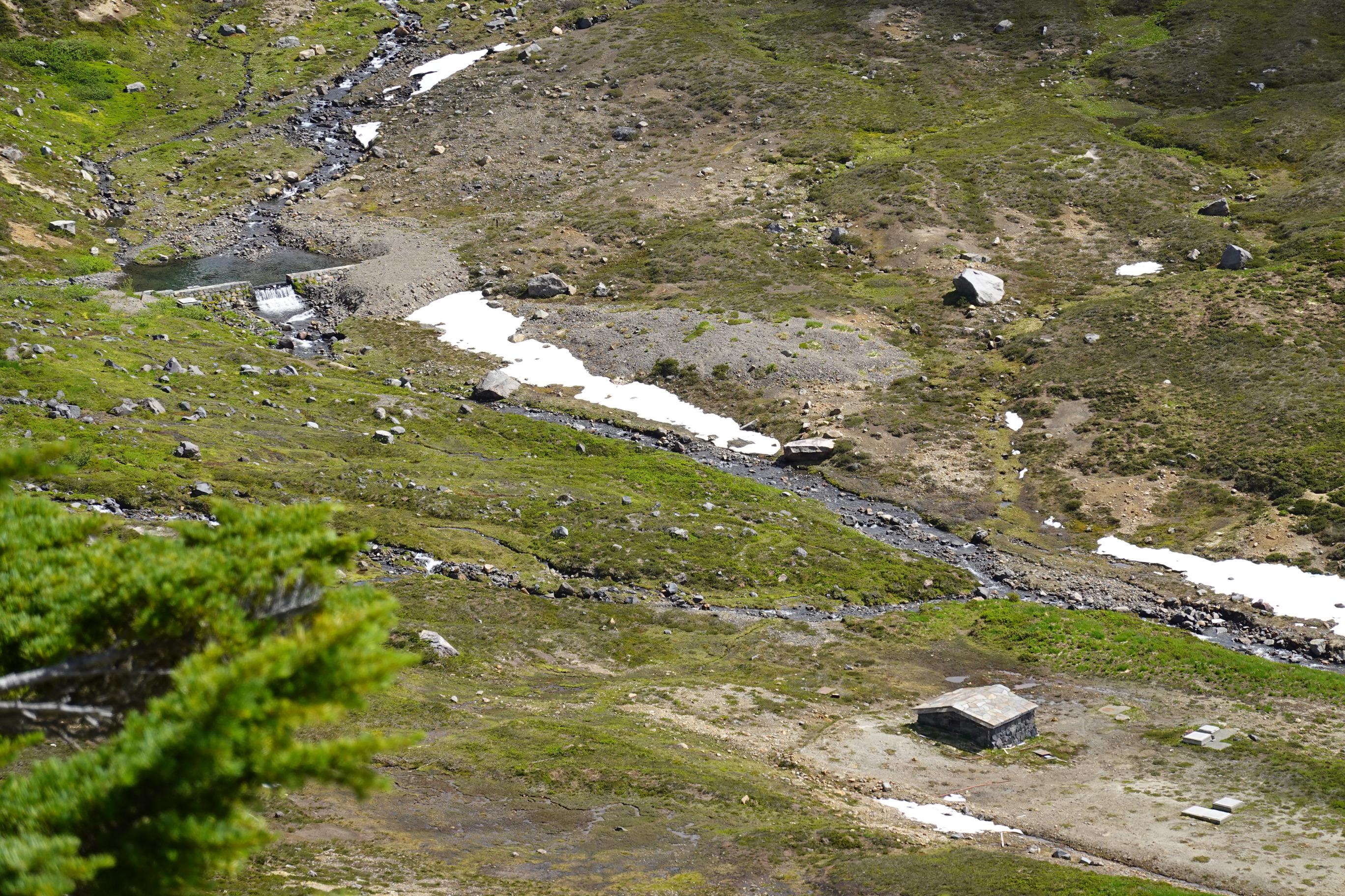 A small stone building next to a creek at the bottom of a wide valley covered in subalpine meadows. 