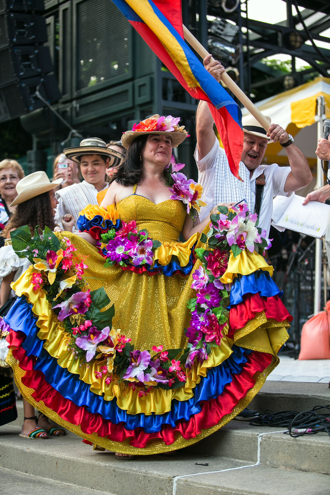 A woman has the long skirt of her golden sequin dress grasped in her hands. The dress is in the horizontal band colors of the Colombian flag: yellow, blue, and red, with a golden-yellow being the most prominent. The dress is adorned with flowers, and so is her wide brim hat. In her group is a man with a wide brim hat waving the Colombian flag.