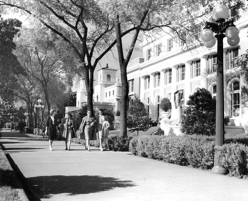 Black & white photograph, Bathhouse Row; sidewalk with women walking on left, Ozark Bathhouse above, Buckstaff Bathhouse on right; light pole along right side.